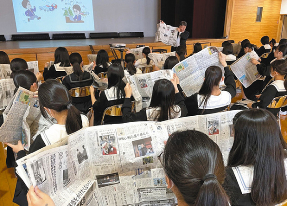 新聞を広げながら鬼頭コーディネーター(奥)の話を聞く生徒たち=瑞穂区の名古屋葵大学中で