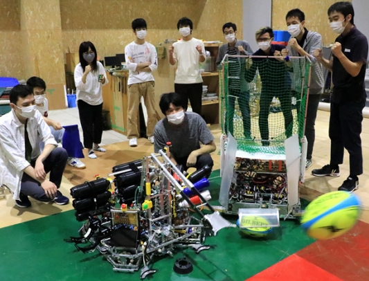 開発したロボット「スコルピウス」がラグビーボール（右下）を蹴り上げる様子を見守るチーム・ロボコンのメンバーたち。右側のロボットは「由美子」＝野々市市扇が丘で