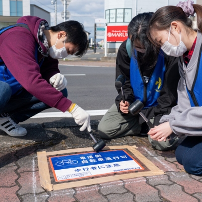 自歩道のシールを圧着させる学生たち＝鈴鹿市白子駅前で