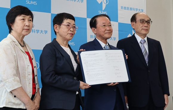 協定書を掲げる井後会長（左から２人目）と高木理事長（同３人目）＝鈴鹿市の鈴鹿医療科学大で