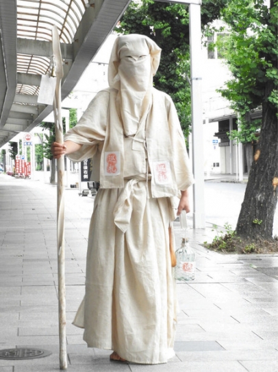 巡礼者のいでたちで「行進する難民」を表現するパフォーマンスアートを行う大西さん＝中区門前町で